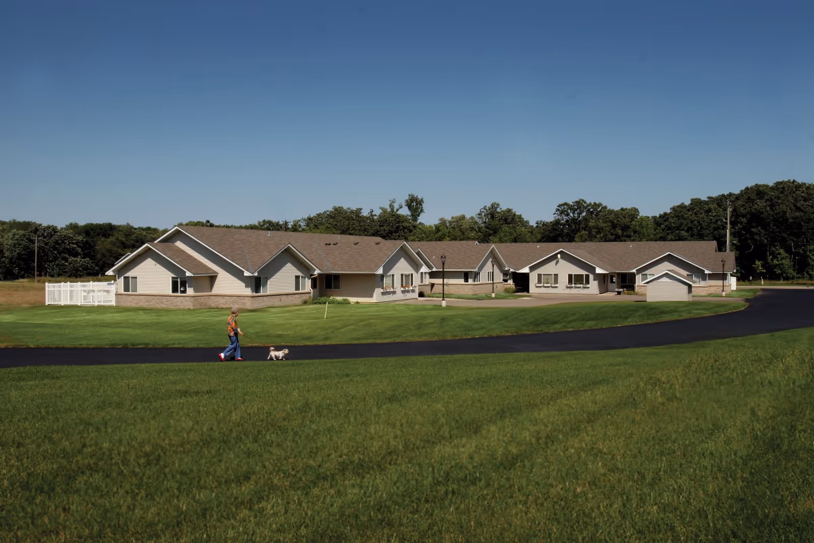Single-story beige senior living building with a wide green lawn and a person walking a small dog along the driveway in front.