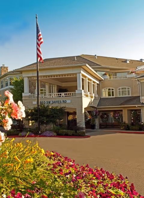 Front exterior of Hearthstone at Murrayhill building with flag, entrance, and blooming flower beds.
