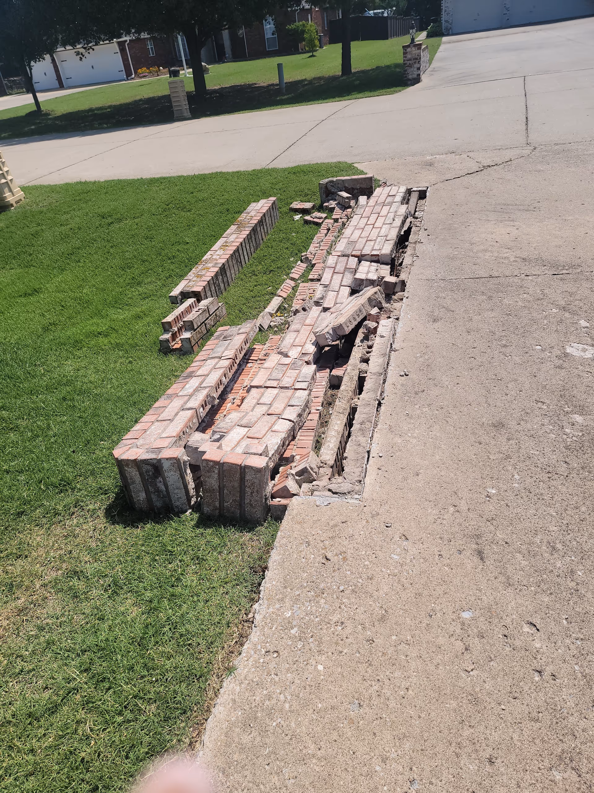 Damaged brick walkway along the grassy edge beside a concrete driveway.