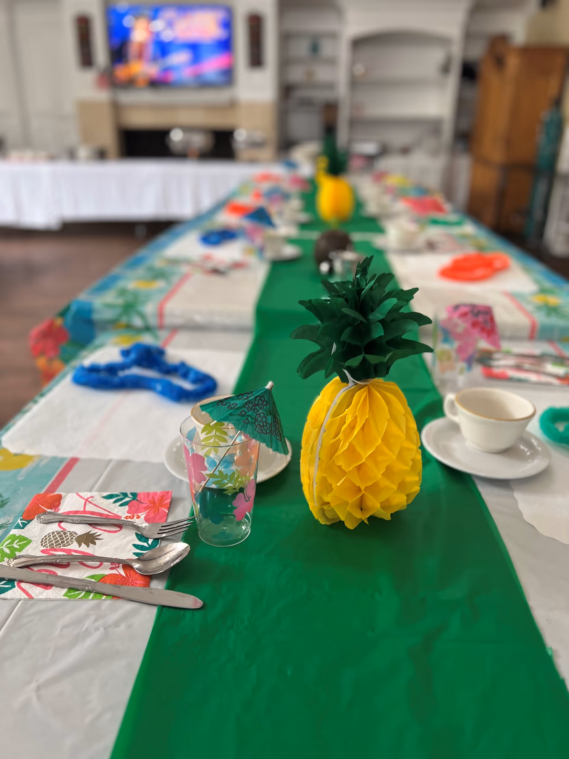 A long dining table decorated with a green table runner and tropical-themed items including a yellow paper pineapple centerpiece, floral napkins, a glass with a small green paper umbrella, and white teacups on saucers. The background shows a blurred television and shelves in a room.