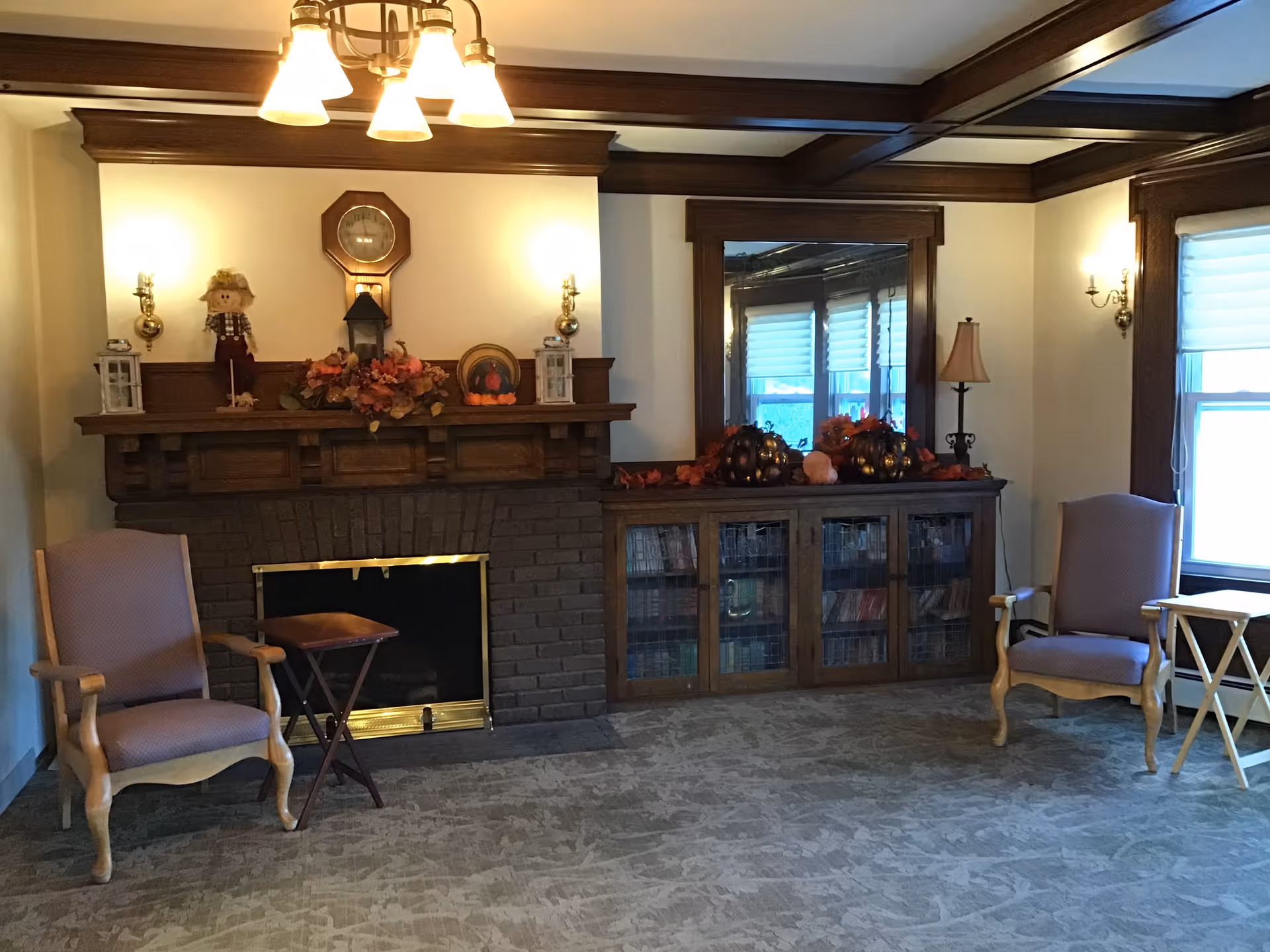 A cozy living room with a dark brick fireplace and wooden mantel decorated with autumn-themed items including a scarecrow, lanterns, and fall foliage. Above the mantel is a wooden clock. On either side of the fireplace are two upholstered armchairs with wooden frames. To the right of the fireplace is a wooden cabinet with glass doors filled with books and decorated with pumpkins and fall leaves. A large mirror hangs above the cabinet, reflecting windows with white blinds. The room has beige walls, carpeted floor, and wooden ceiling beams. A chandelier with five lights hangs from the ceiling.