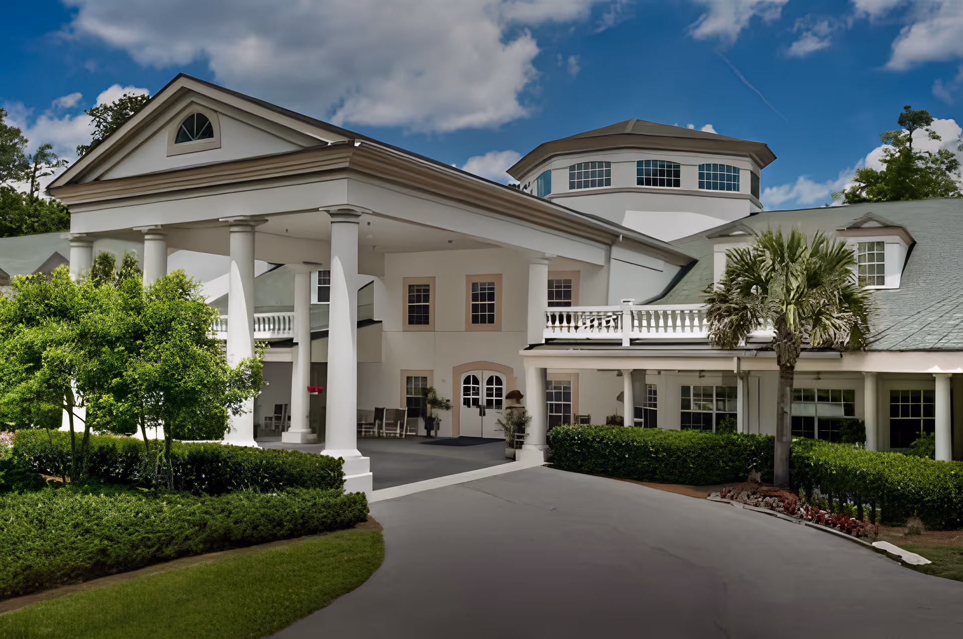 Front exterior view of a large senior living facility building with white columns, a covered entrance, multiple windows, and surrounding greenery including bushes, trees, and a palm tree under a blue sky with scattered clouds.