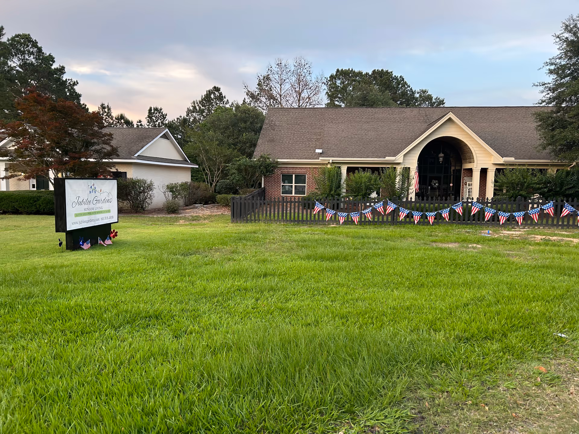 Front view of Jubilee Gardens Senior Living facility with a fenced lawn, sign, and flags.