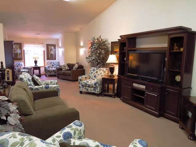 A spacious living room with multiple upholstered chairs and sofas arranged around a large wooden entertainment center holding a flat-screen TV. The room is warmly lit with table lamps and wall sconces, and decorated with framed artwork and a large potted plant.
