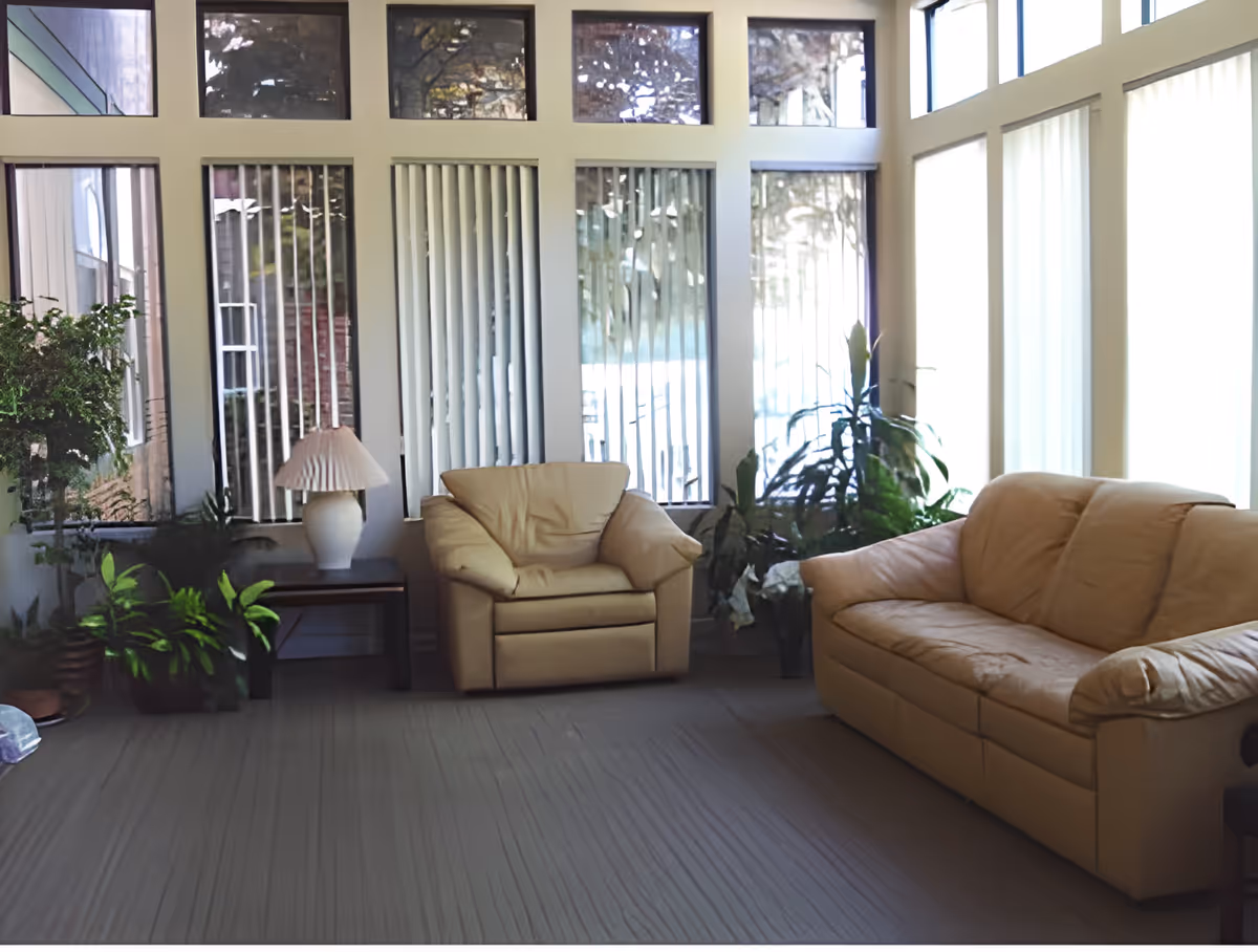 A bright living room with large windows covered by vertical blinds, beige leather armchair and sofa, a wooden side table with a white lamp, and several green potted plants.