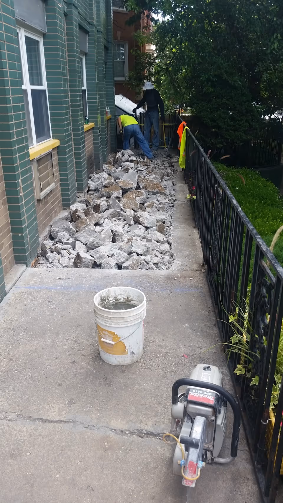 Exterior of a building with rubble, workers, and a bucket on the sidewalk.