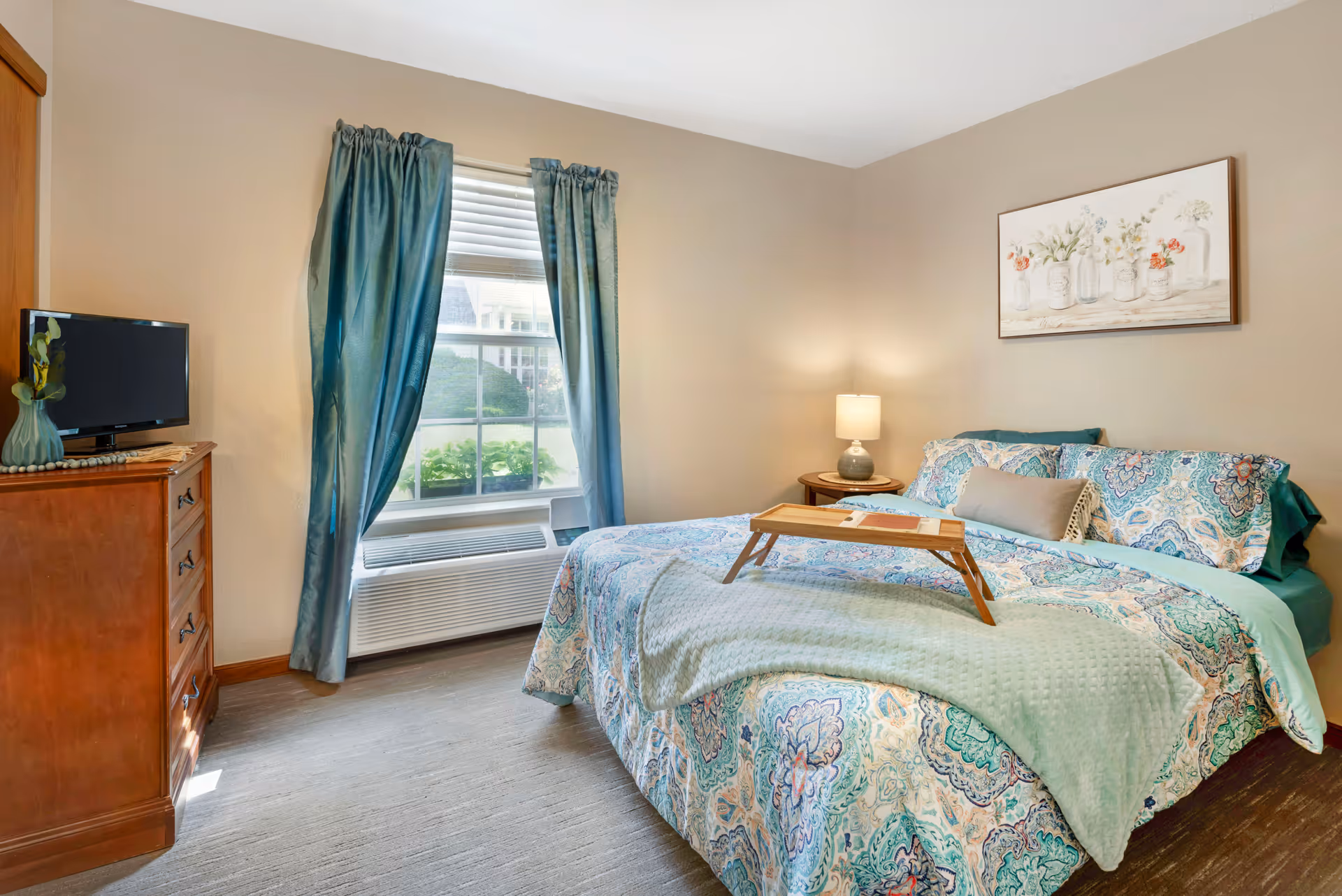 A cozy bedroom with a bed covered in a colorful patterned comforter and pillows. A wooden tray is placed on the bed. There is a nightstand with a lamp beside the bed and a wooden dresser with a small TV and a decorative vase. Blue curtains frame a window letting in natural light.