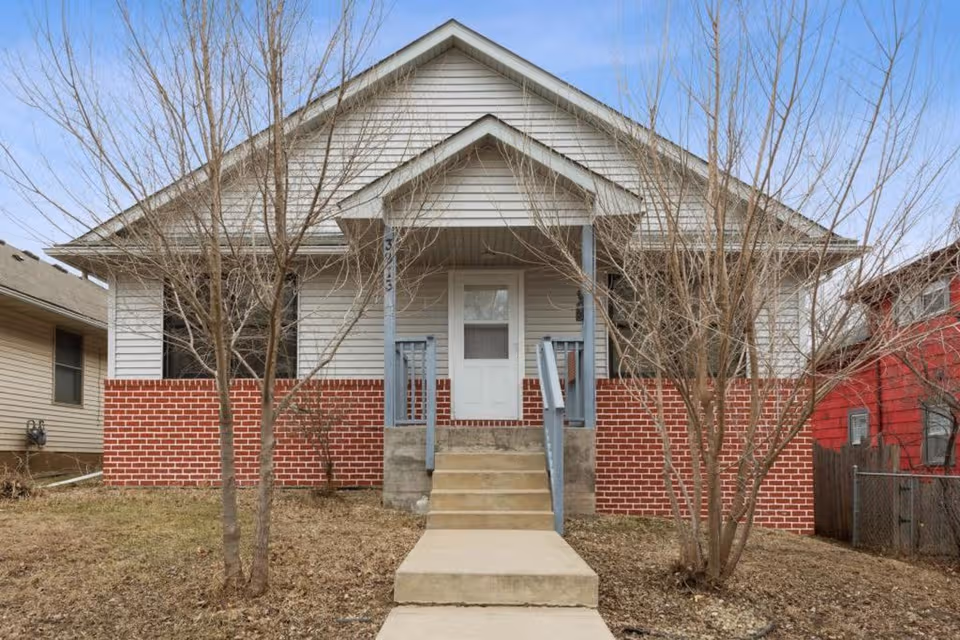 Front view of a small brick-and-siding house with a porch, steps, and leafless trees.