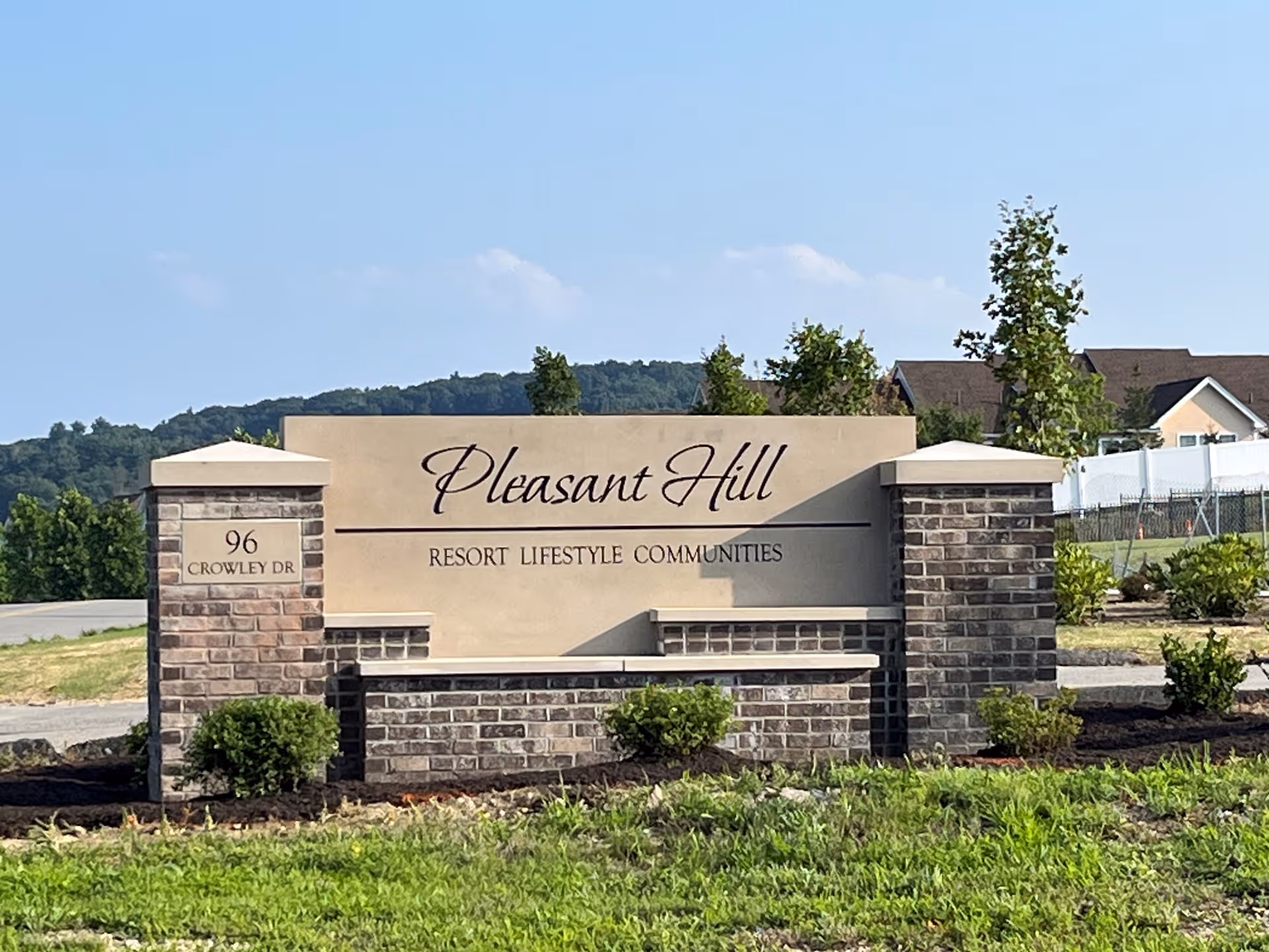 A large stone and brick sign at the entrance of Pleasant Hill Resort Lifestyle Communities with the address 96 Crowley Dr, surrounded by grass and small bushes, with houses and trees in the background under a clear blue sky.