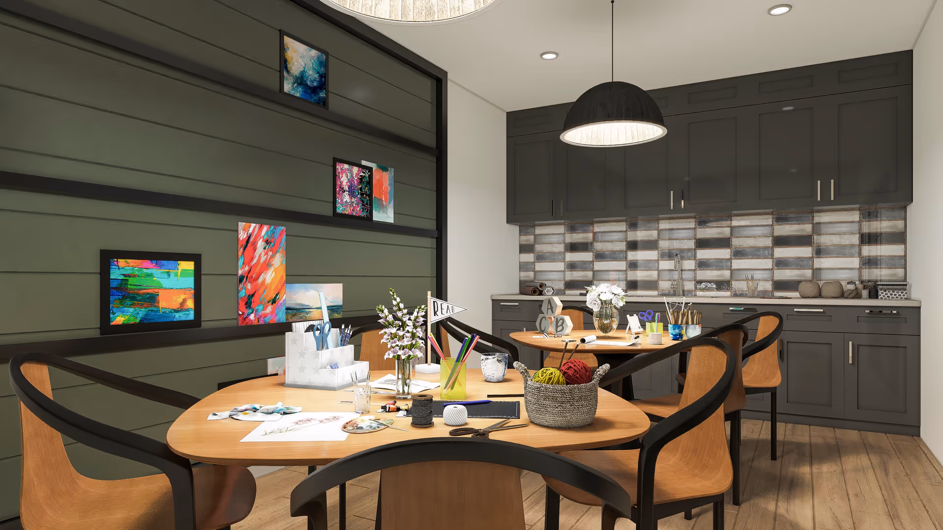 An activity room with round wooden tables and chairs covered in craft supplies, set against a kitchenette with gray cabinets and a tiled backsplash.
