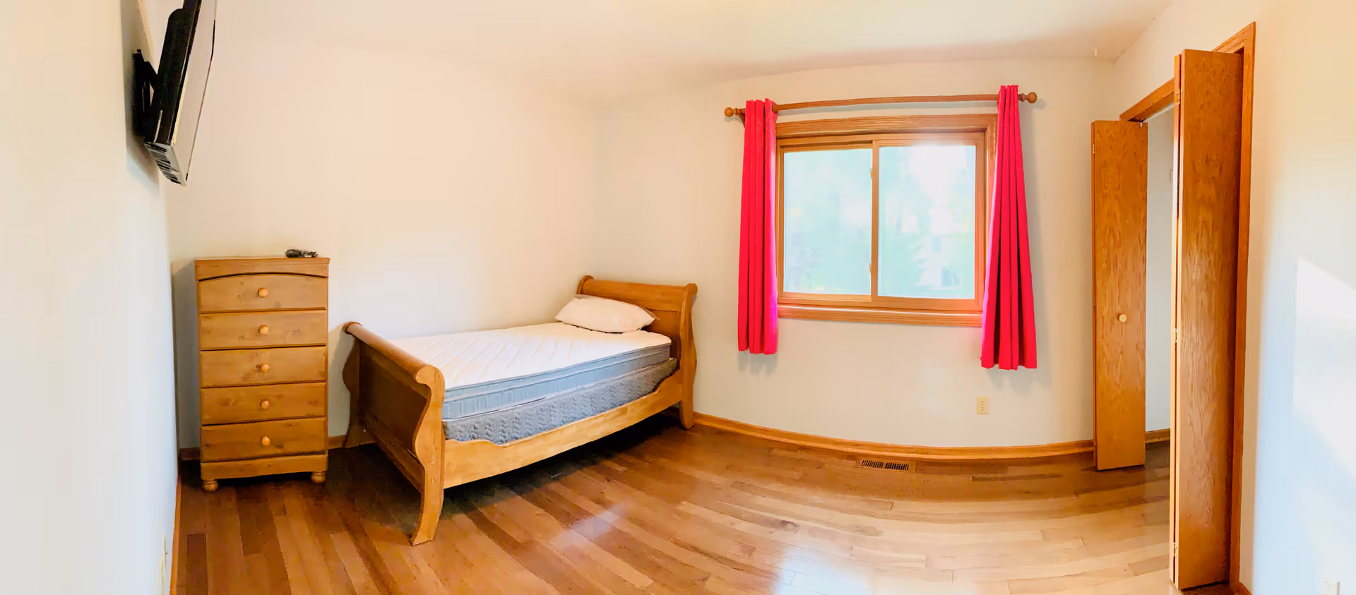 A simple bedroom in a senior living facility with a bed, dresser, window, and pink curtains.