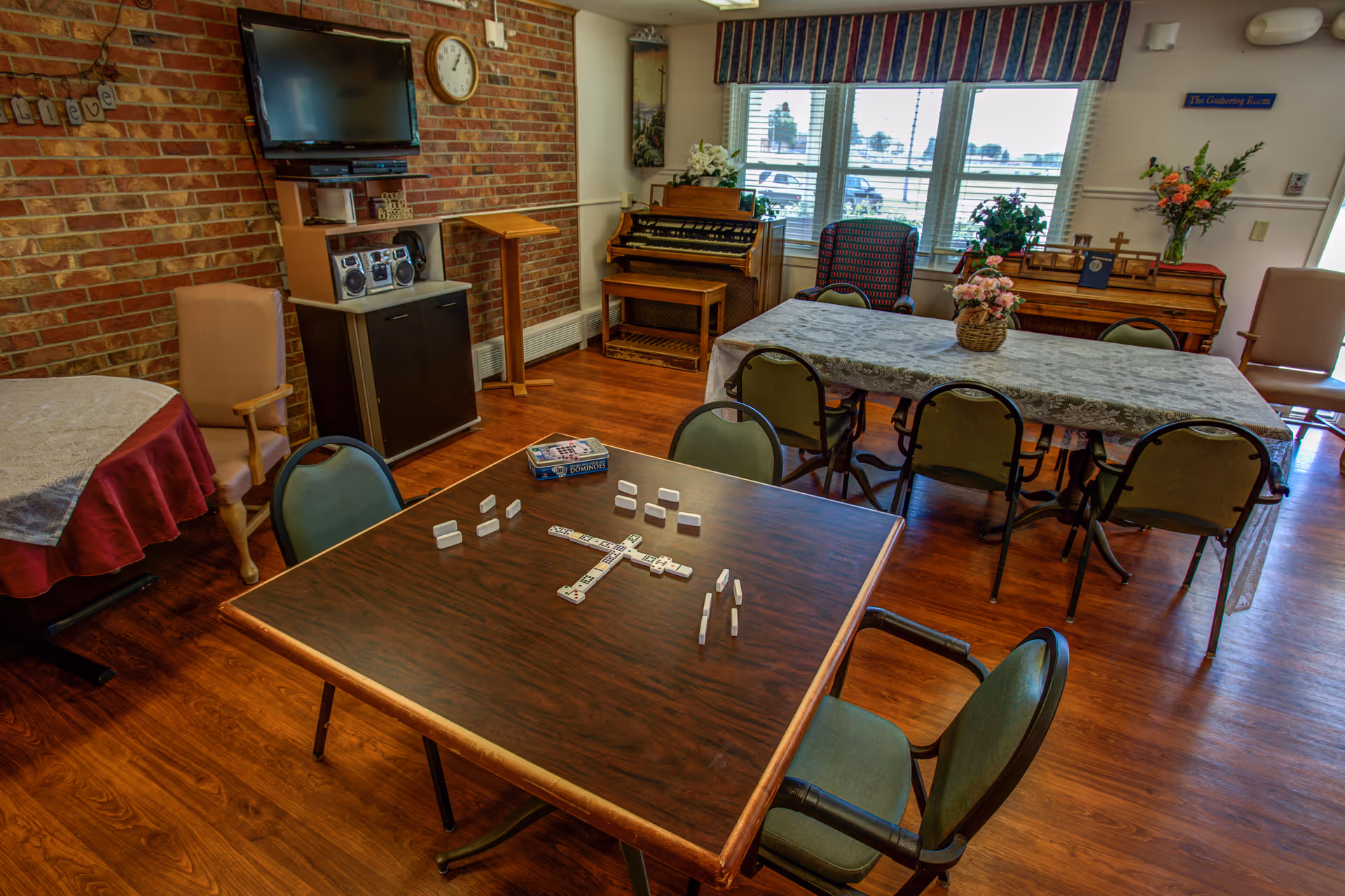 Interior view of a senior living common room with tables, chairs, and a domino setup.