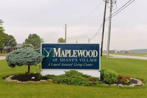 A Maplewood of Shane's Village sign sits on a grassy lawn with shrubs.