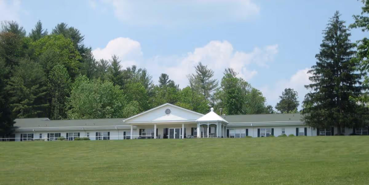 Long single-story white health care building with a central entrance and gazebo set behind a wide grassy lawn and trees.