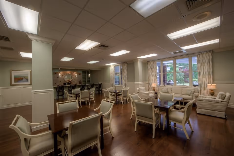 A spacious and well-lit common area with multiple tables and chairs arranged for seating. There are comfortable sofas and armchairs near large windows with curtains, allowing natural light to enter. The room has a warm wooden floor, soft lighting from ceiling fixtures, and a decorative wall with a mirror and flowers in the background.