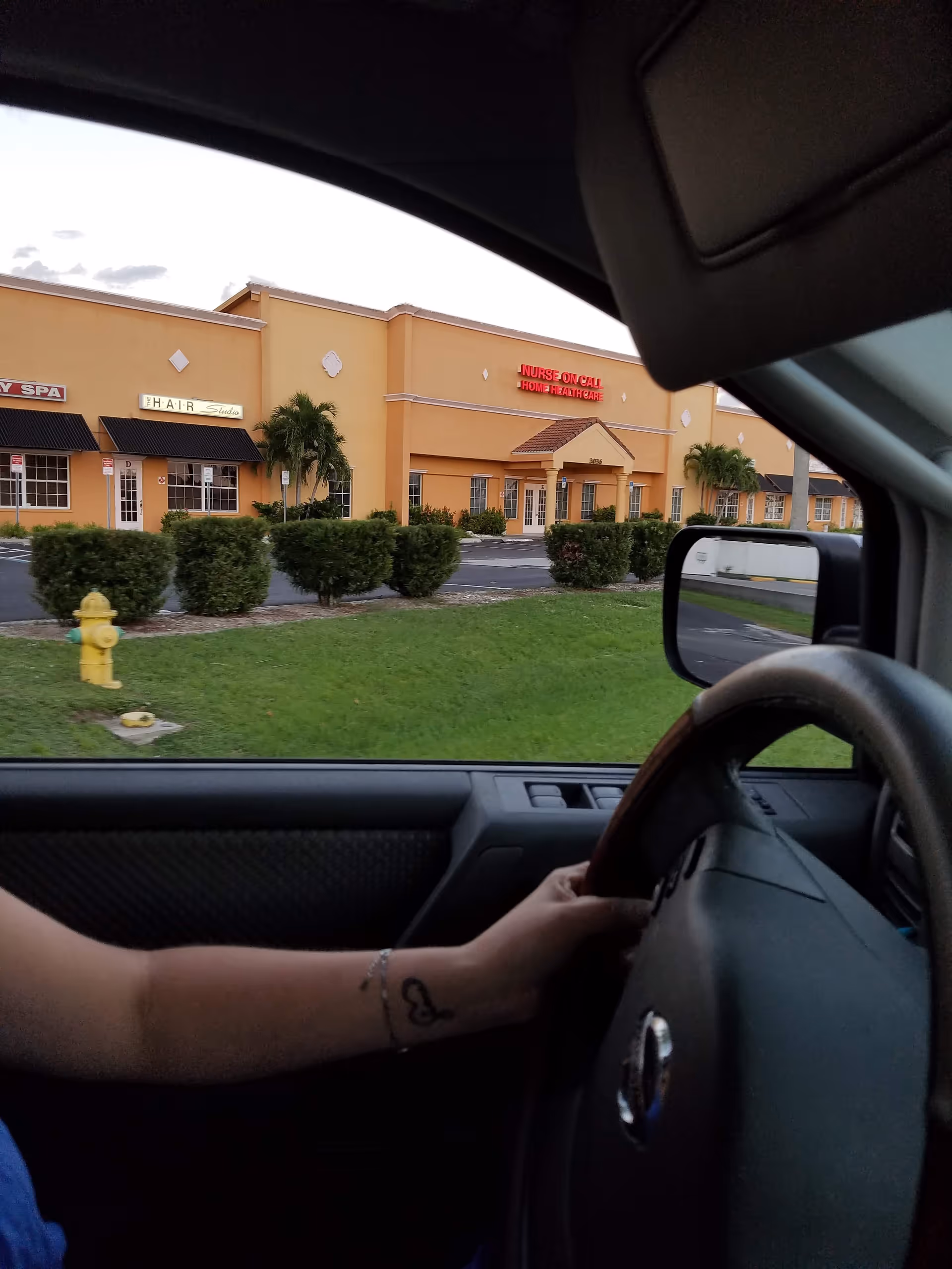 Driver's arm and steering wheel in foreground; orange storefronts across parking lot