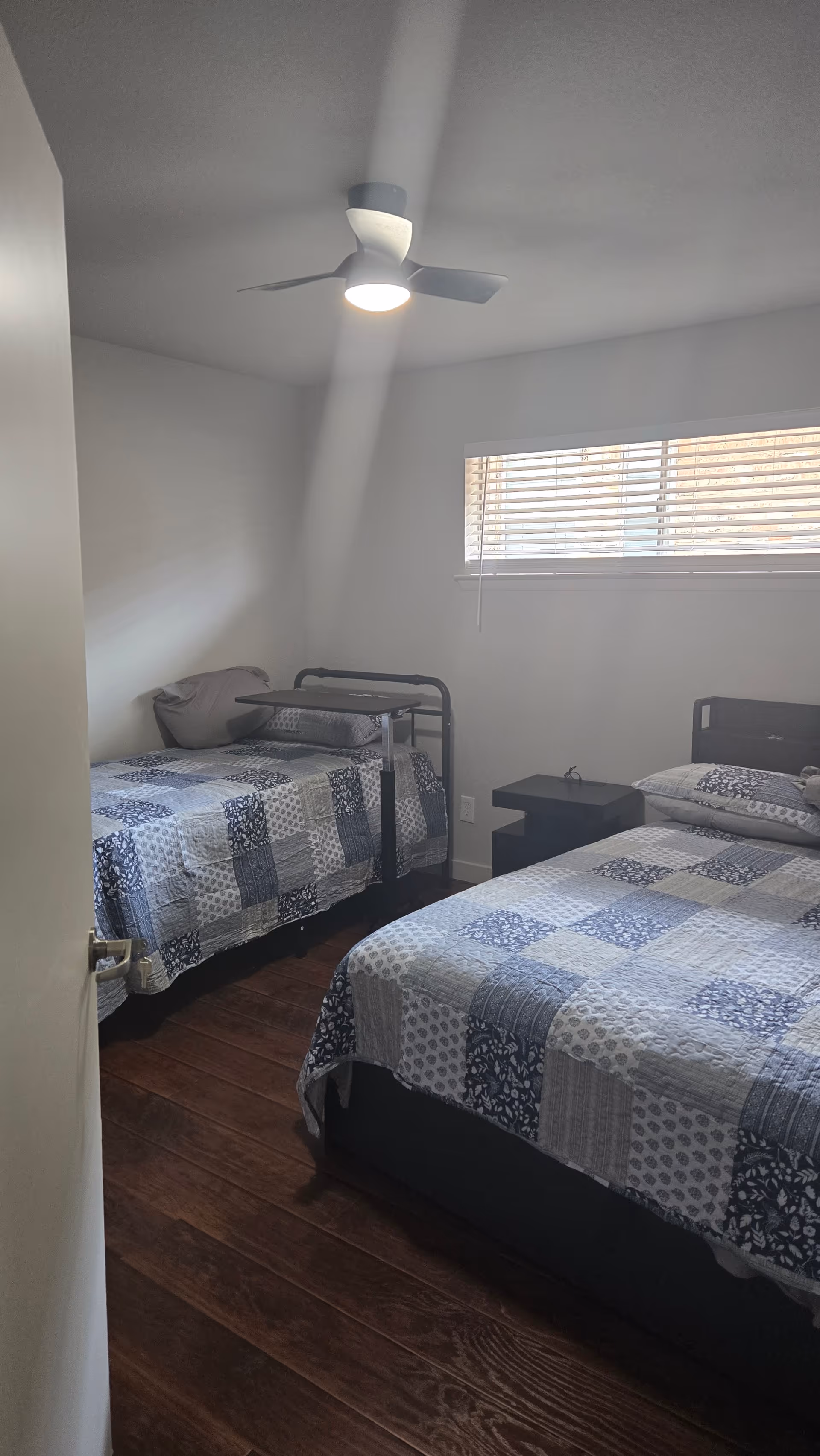 Two twin beds with patchwork blue quilts, a nightstand, window blinds, and ceiling fan.
