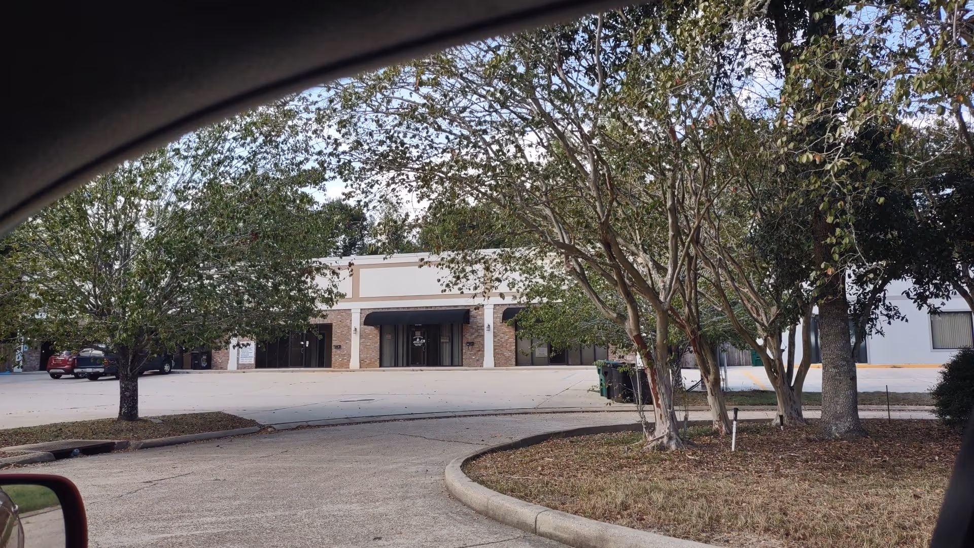 Facade of a beige storefront-style building with brick accents and trees in a parking lot