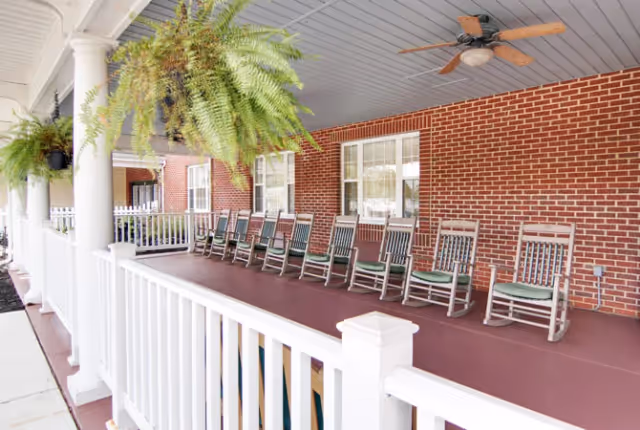 Covered front porch with a row of wooden rocking chairs, hanging ferns, a ceiling fan, and white railing against a red brick wall.