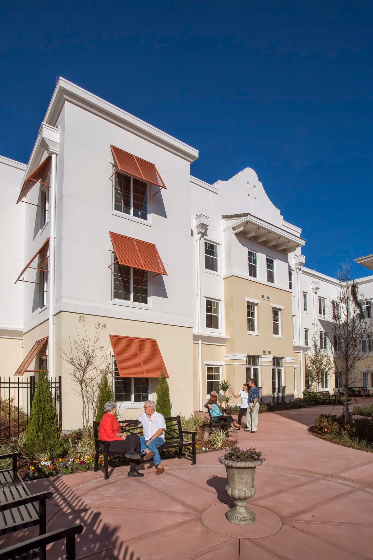 Courtyard of a light-colored assisted living building with residents sitting on benches and walking along a paved path under a clear blue sky.