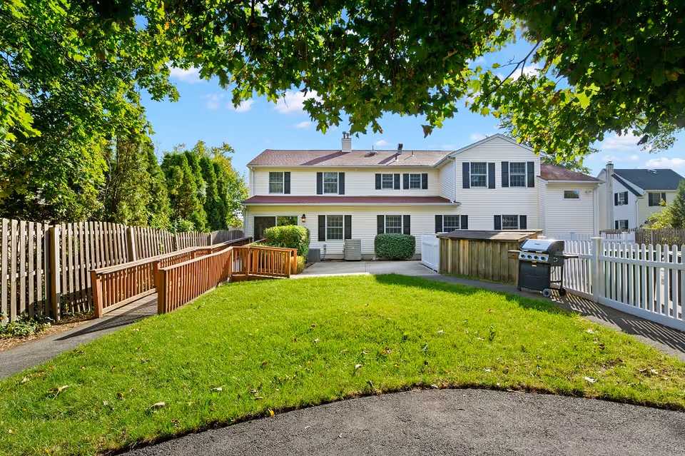 Two-story white house with green lawn, fenced backyard, and wooden ramp under trees.
