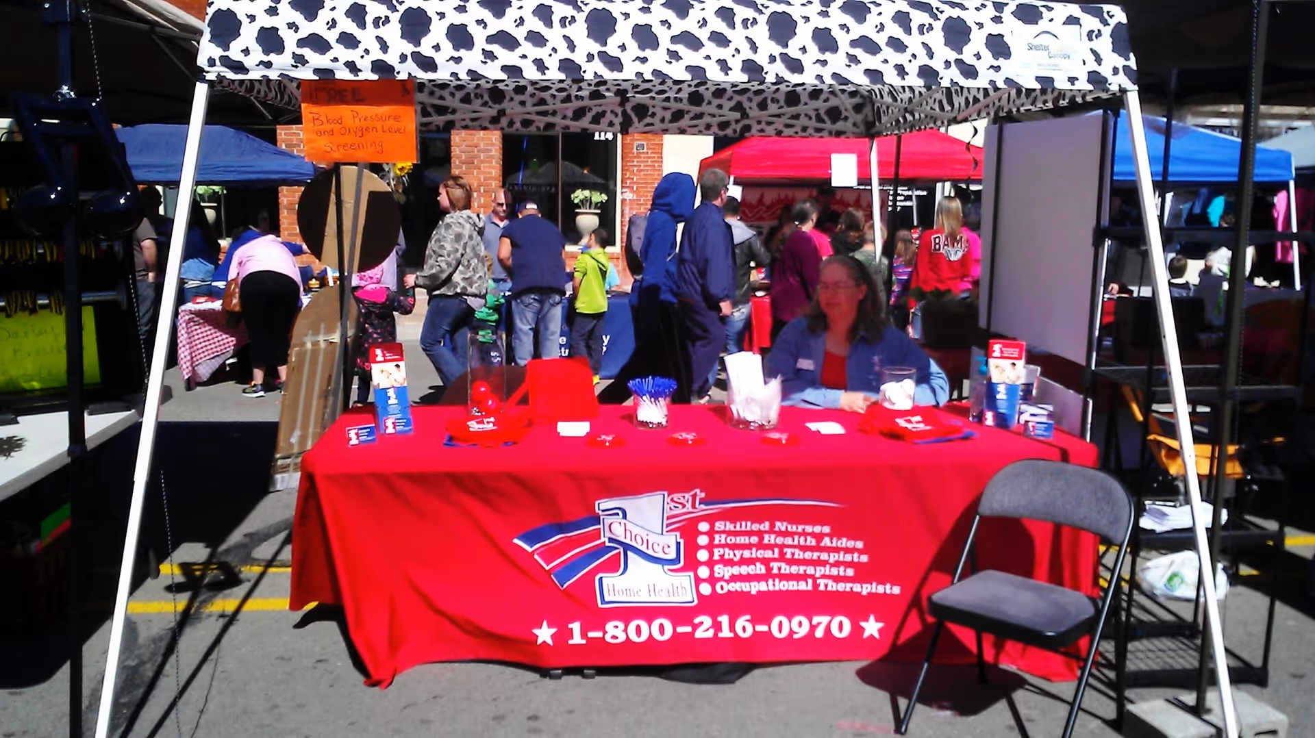 An outdoor information booth with a red tablecloth promoting First Choice Home Health, with people nearby.