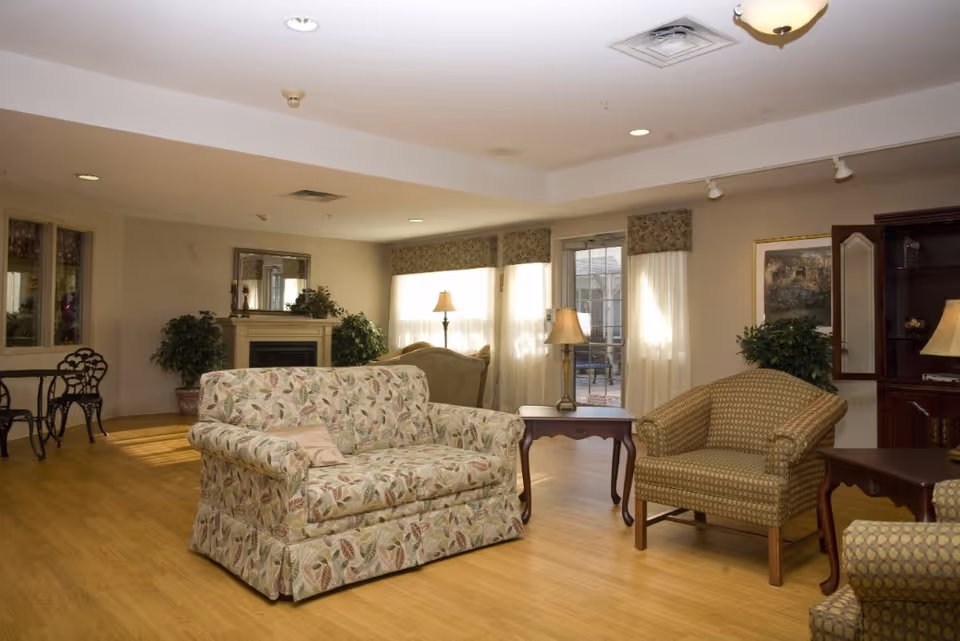 A cozy living room area in a senior living facility with a floral patterned loveseat, two armchairs, wooden side tables with lamps, a fireplace with a mirror above it, potted plants, and large windows with curtains allowing natural light to fill the room.