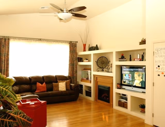 Bright living room with a brown leather sofa, built-in shelves and fireplace, TV, ceiling fan, and hardwood floors.