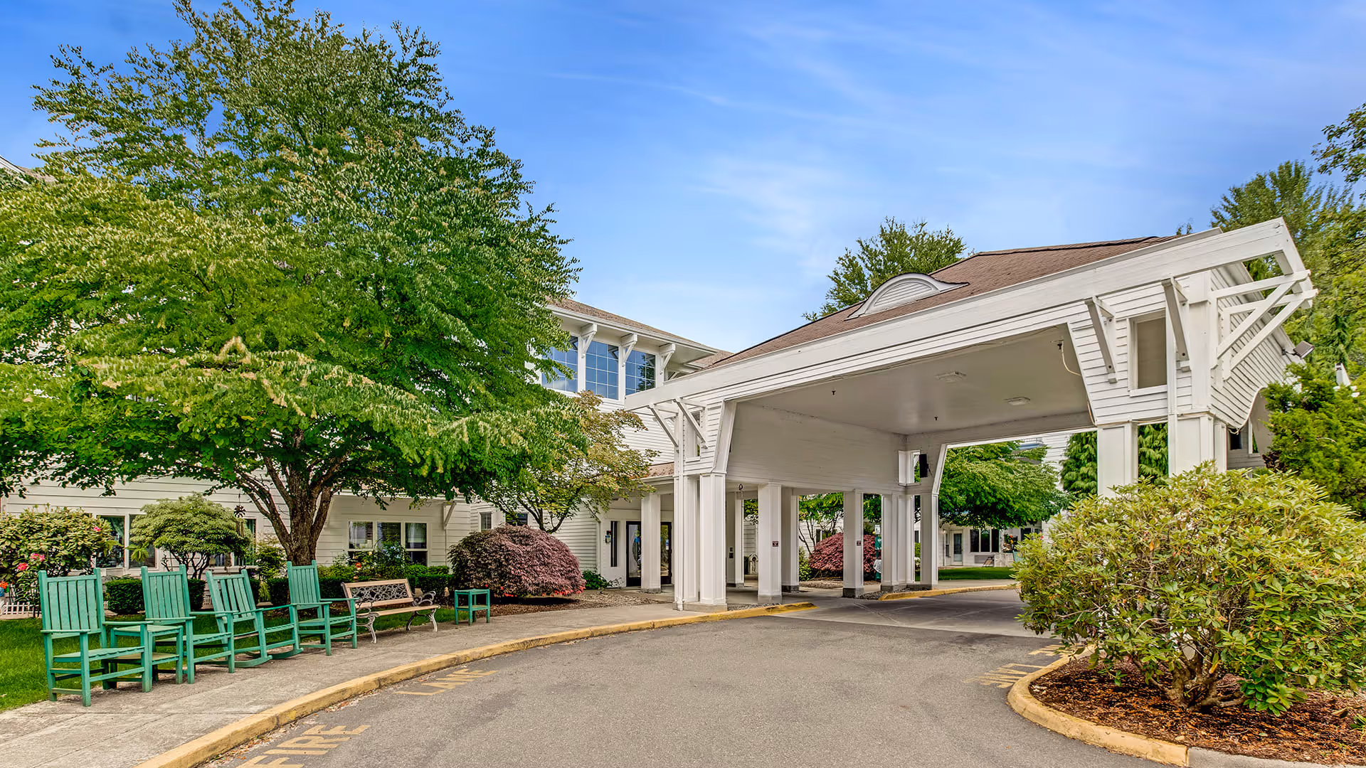 Entrance driveway of Holiday South Hill facility with a covered drop-off area, surrounded by green trees, bushes, and a row of green rocking chairs and a bench along the sidewalk.