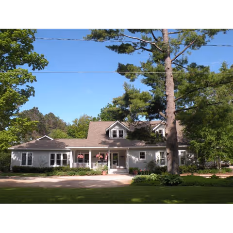 Front exterior of a single-story house with a covered porch, hanging flags, and large trees on a sunny day.