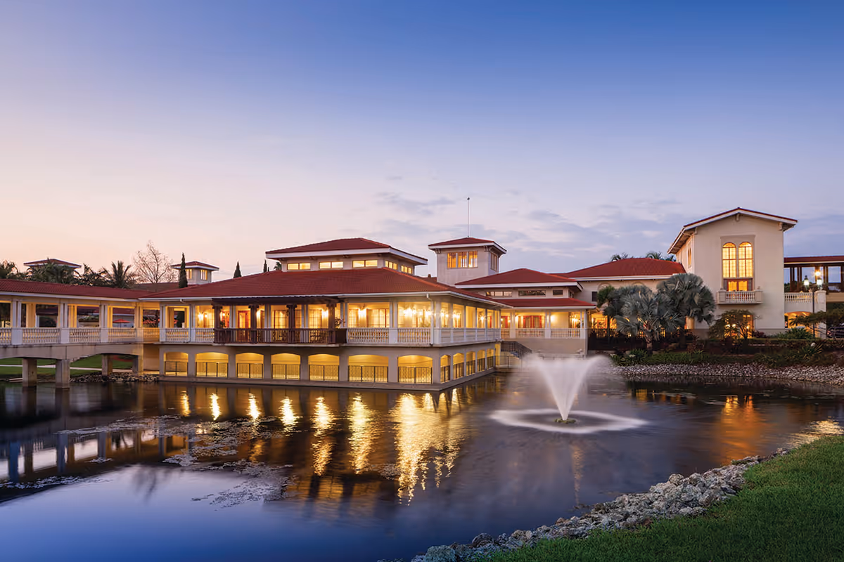A large senior living facility building with red roofs and multiple windows, situated beside a pond with a water fountain in the center. The building is illuminated with warm lights during dusk, reflecting beautifully on the water surface. There is a grassy area and rocks along the pond's edge.