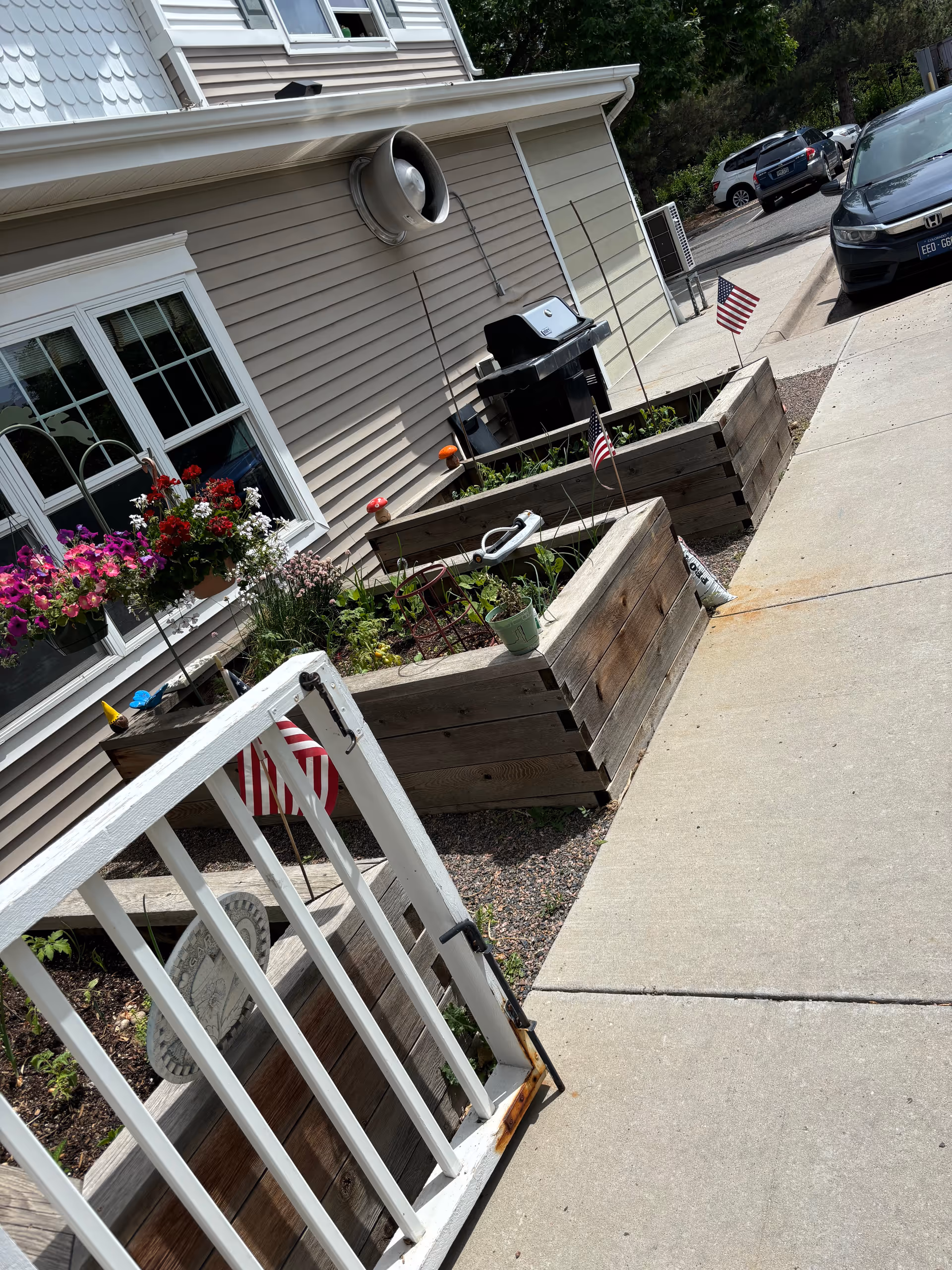 Front exterior of a senior living building with raised wooden planters, a white gate, American flags, and a sidewalk.