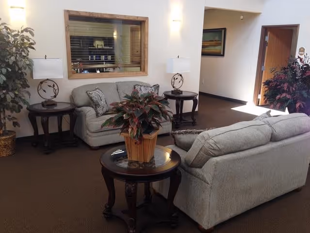 A cozy living room area with two light gray sofas arranged around a round wooden coffee table with a potted plant. There are two matching side tables with decorative lamps on either side of one sofa. A large window with wooden trim is on the wall behind one sofa, and a hallway with a framed picture is visible in the background.