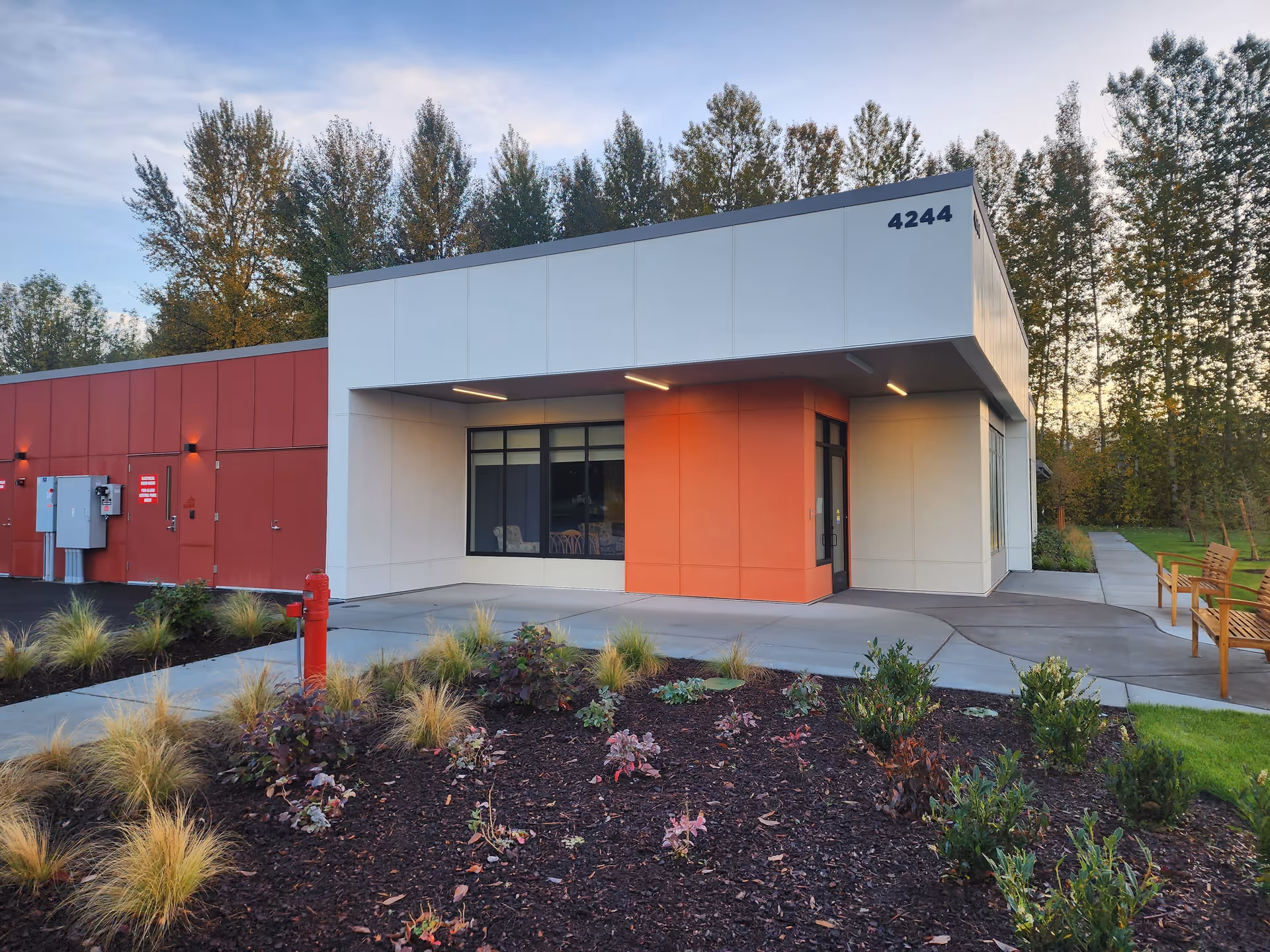 Modern senior living facility exterior with a white and orange front entrance, benches, and landscaping.