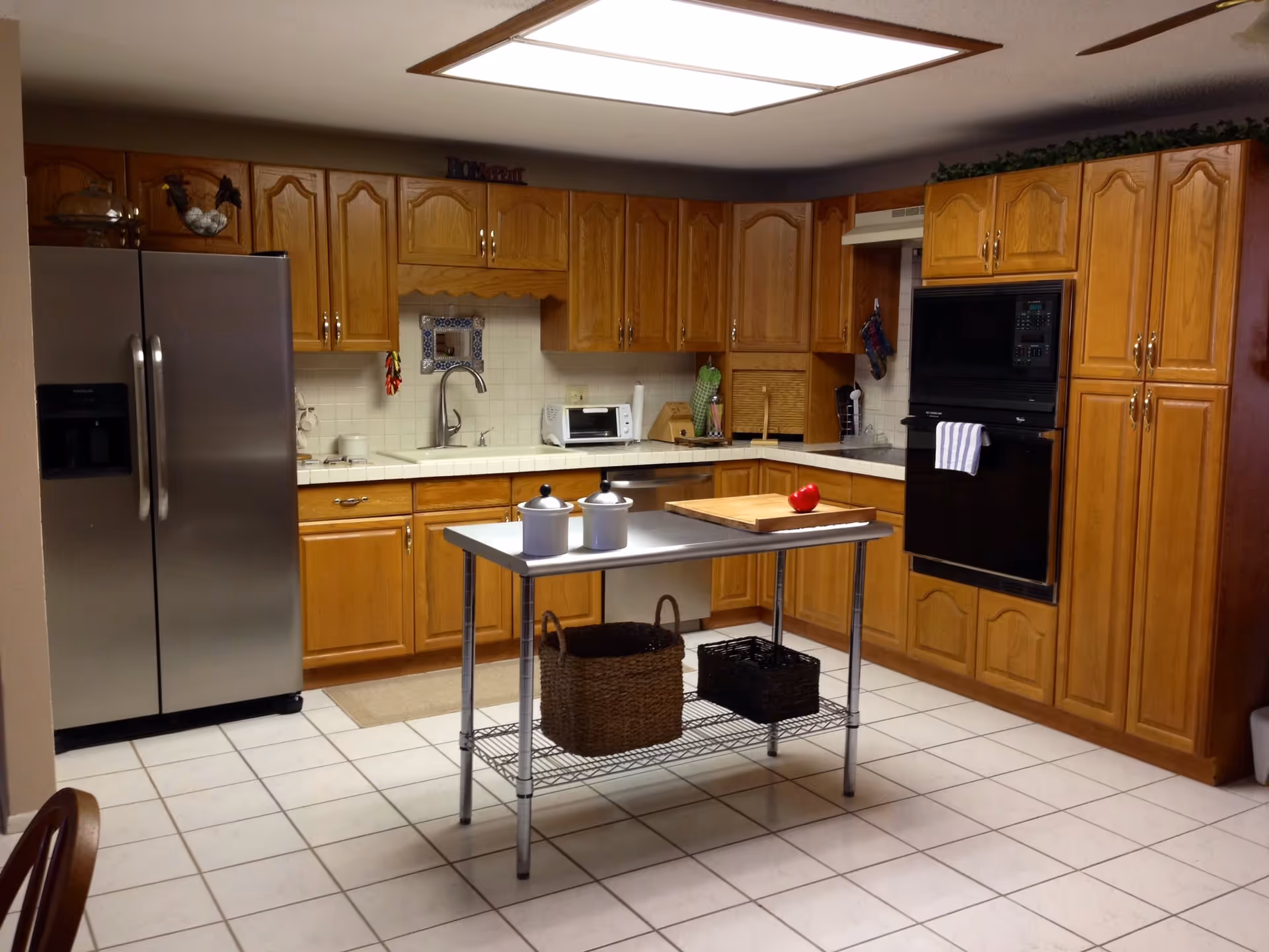 Bright kitchen with oak cabinets, a stainless refrigerator, a metal center island and tiled floor.