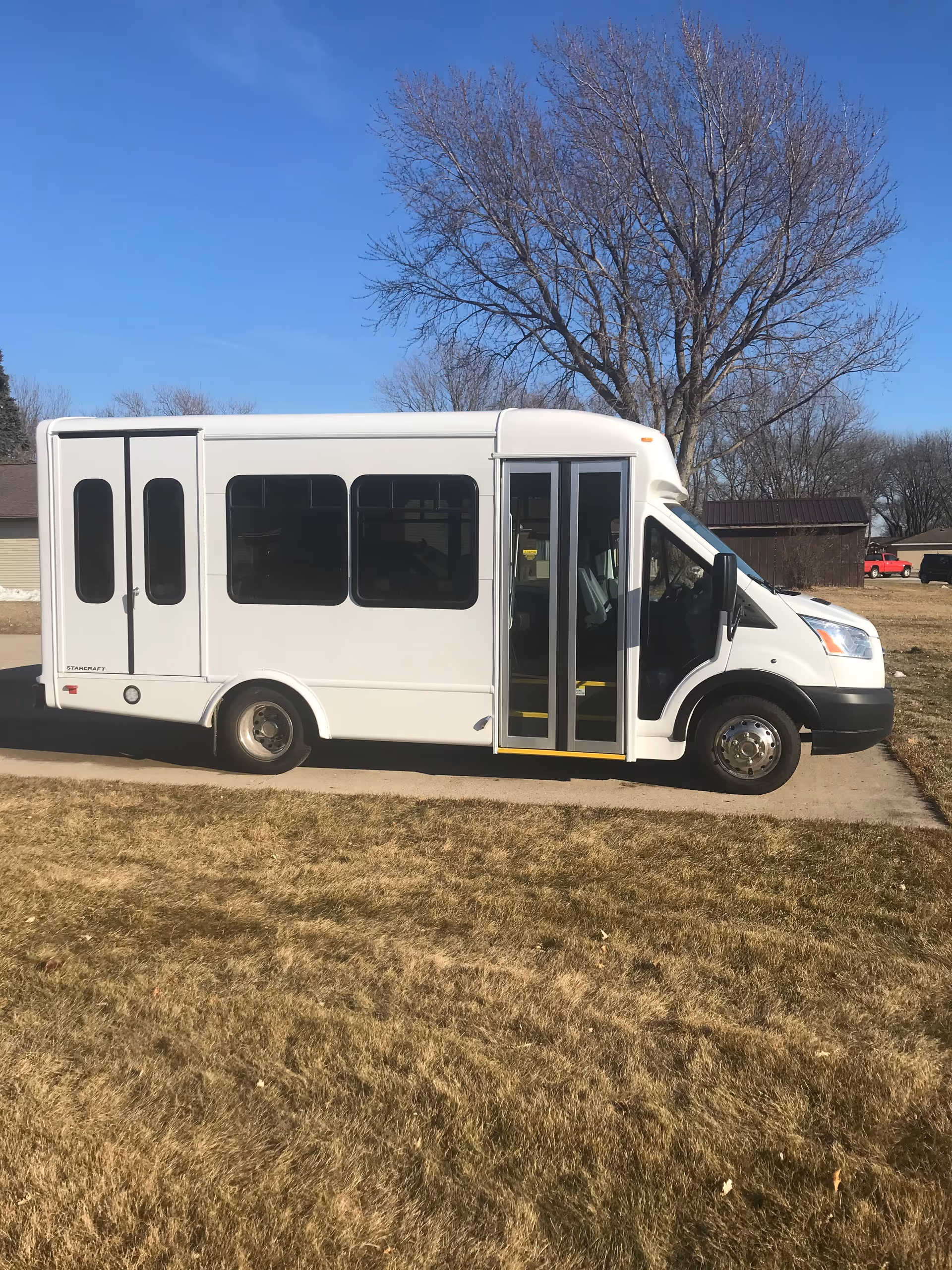 A white passenger shuttle/minibus parked on a driveway beside a grassy yard with a large leafless tree and clear blue sky.