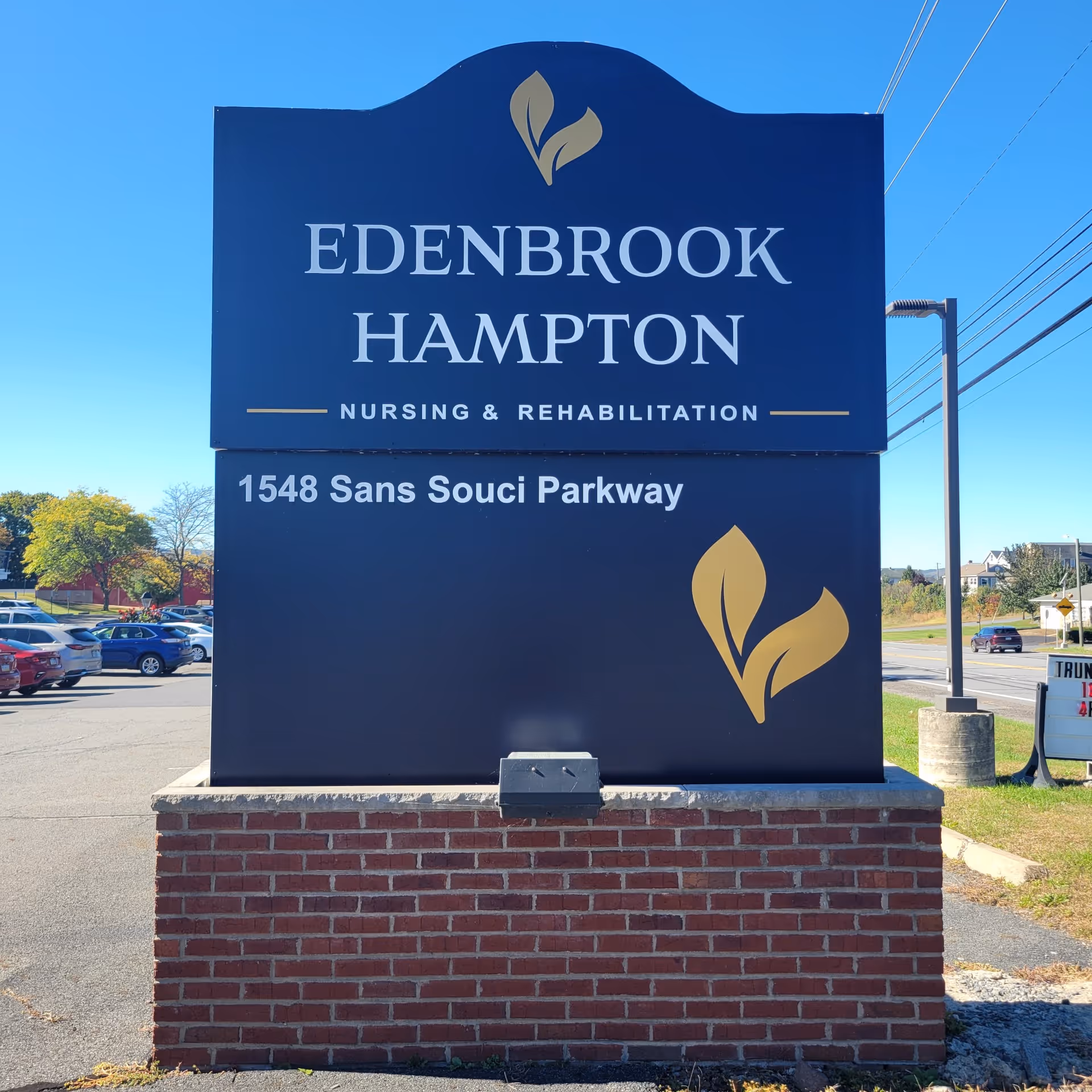 Large blue Edenbrook Hampton Nursing & Rehabilitation sign with brick base near parking.