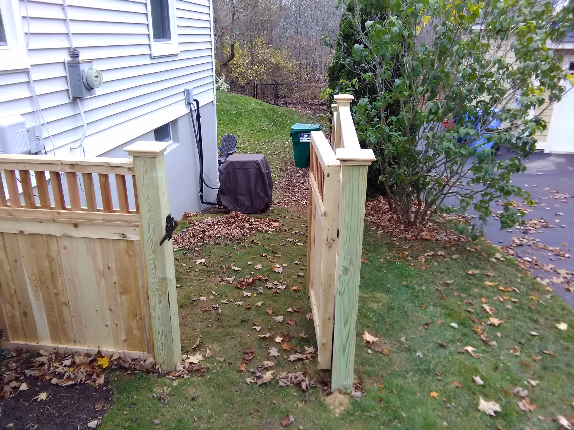 Outdoor backyard with wooden gate, grill, trash bin, and fallen leaves.