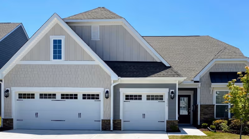 Front exterior of a light-gray house with a three-car garage, driveway, and small lawn under a clear blue sky.