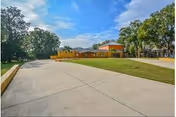 Wide concrete driveway leads to a tan building with orange roof, surrounded by lawn and trees.