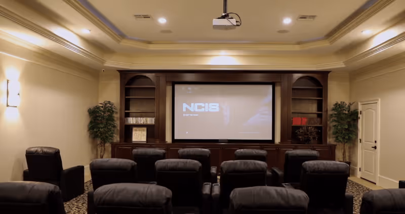 A cozy home theater room with multiple black leather recliner chairs arranged in rows facing a large screen displaying the NCIS show. The room has built-in wooden shelves on either side of the screen, decorative plants in the corners, and soft wall lighting.