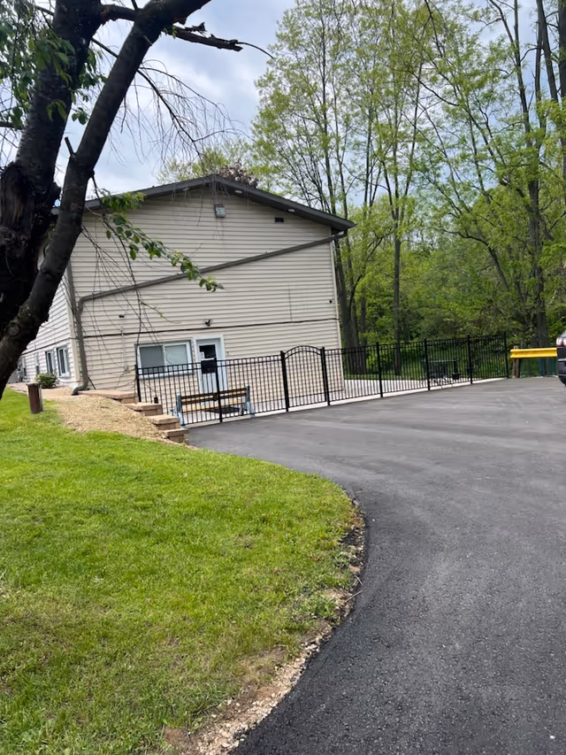 Exterior view of a two-story beige building with a sloped roof, surrounded by green grass and trees. There is a black metal fence enclosing a small patio area with a bench, and a paved driveway curves in front of the building.