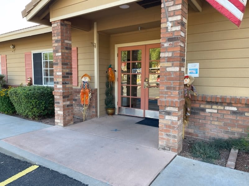 Entrance to a building with double glass doors framed by brick pillars. Two scarecrow decorations are placed on either side of the entrance. There is an American flag hanging above the right side of the entrance. Bushes and windows with pink shutters are visible on the left side of the building.