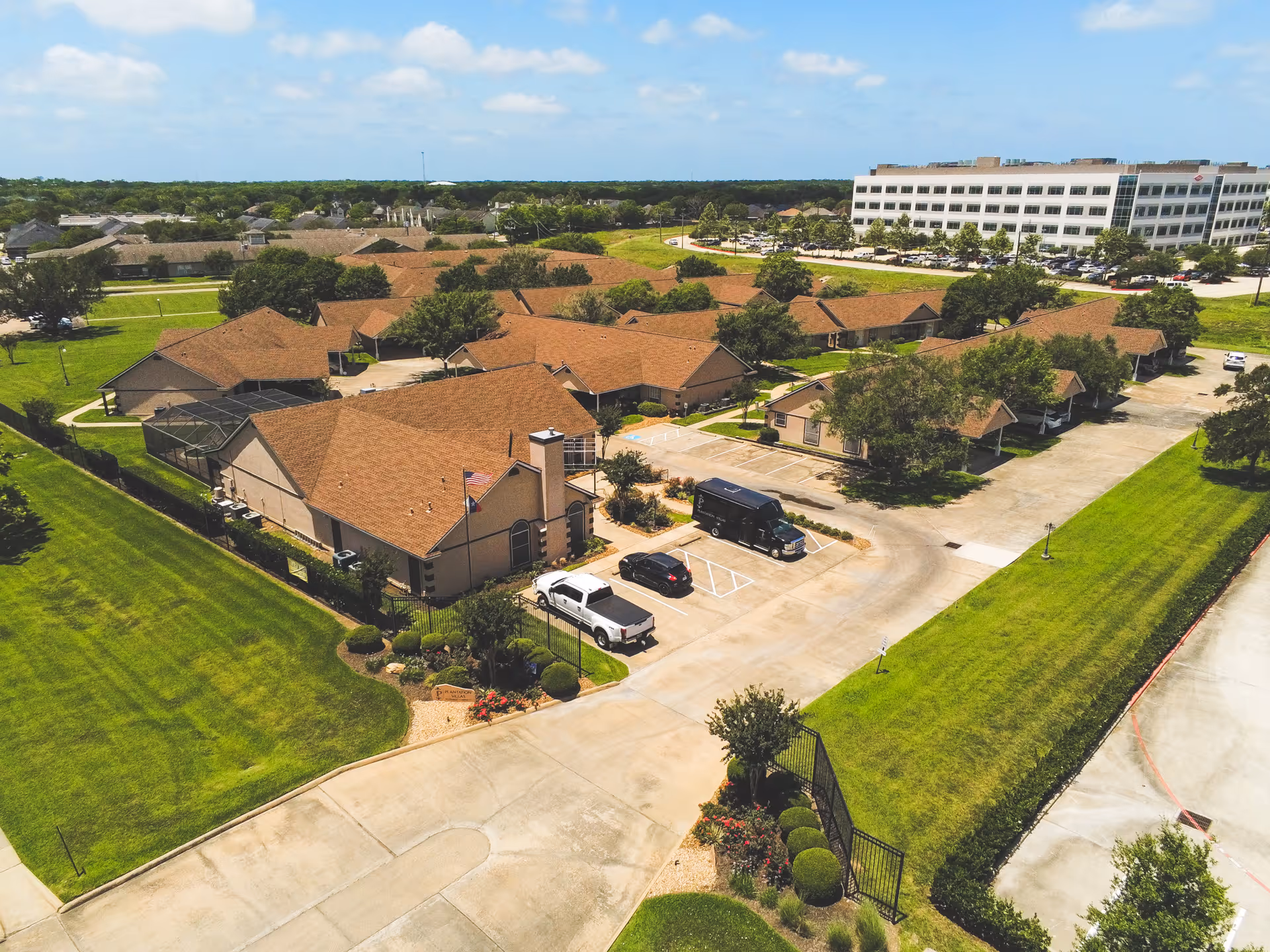 Aerial view of Plantation Villas senior living facility showing multiple single-story buildings with brown roofs, surrounded by green lawns and trees, with a parking area containing several vehicles and a large multi-story building in the background under a partly cloudy sky.