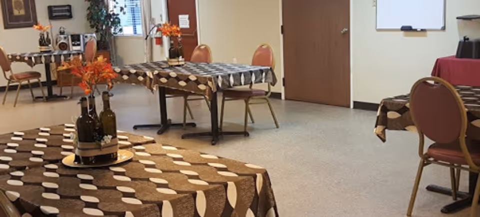 Community dining room with tables covered in brown patterned tablecloths, chairs, and vases of orange flowers.