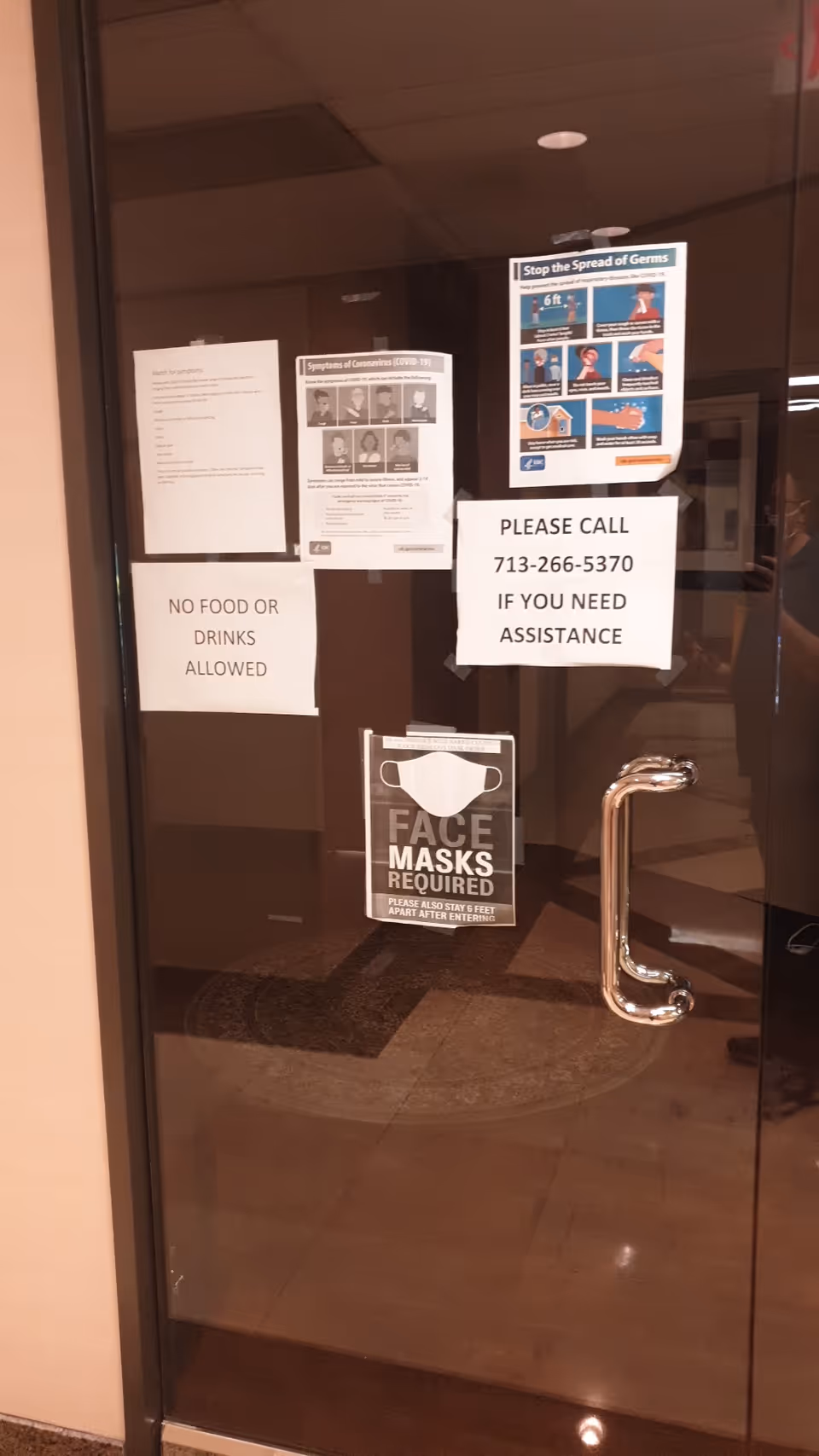 Glass door with multiple posted notices, including masks required and assistance contact in a facility lobby.