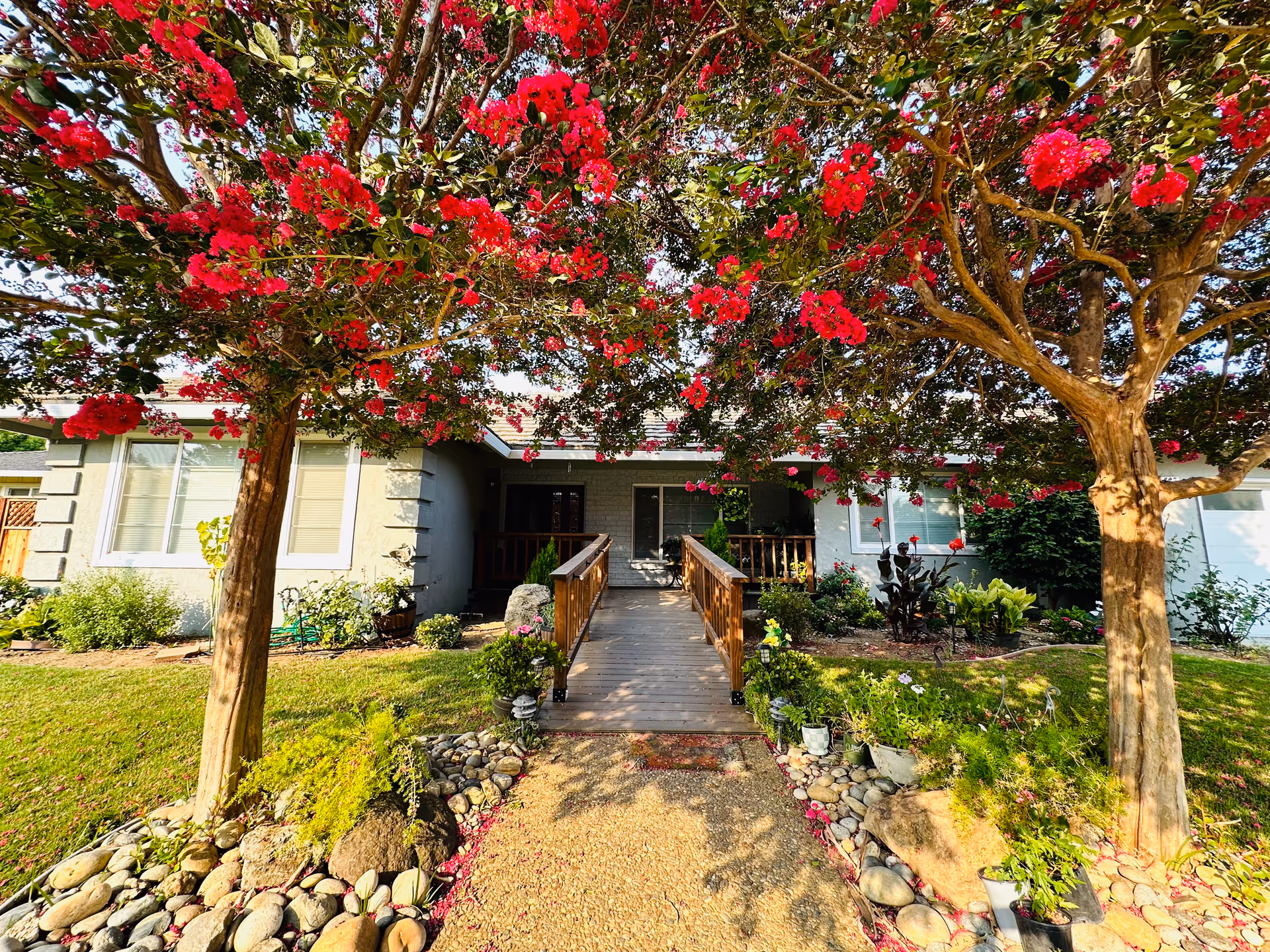 Front entrance of Wimbledon Walnut Creek Care Home with a wooden ramp, blooming trees, and garden.