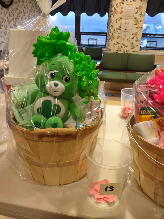 A gift basket wrapped in clear plastic with a large green bow on top. Inside the basket is a green stuffed bear with a white belly featuring a green heart. The basket is placed on a table next to a clear plastic cup labeled with the number 13 containing pink paper slips. In the background, there is a green couch, floral wallpaper, and windows with dark curtains.