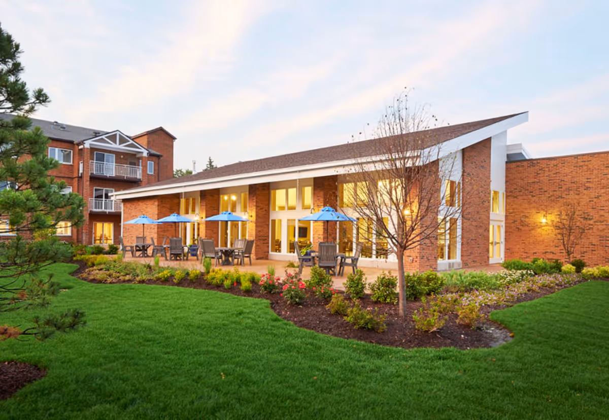 Brick senior living building with a patio area featuring blue umbrellas, outdoor seating, and a landscaped lawn.