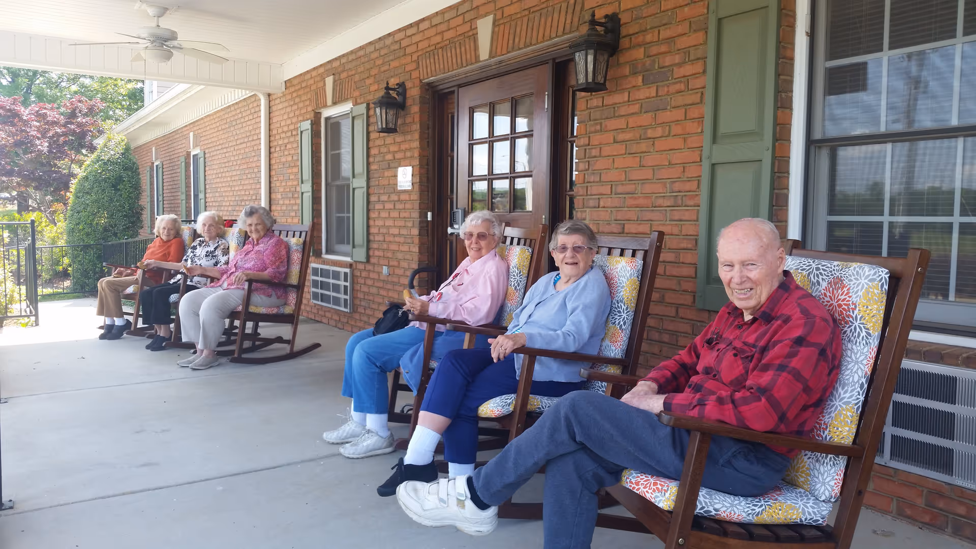 Six elderly individuals sitting on cushioned rocking chairs on a covered porch outside a brick building, enjoying a sunny day.